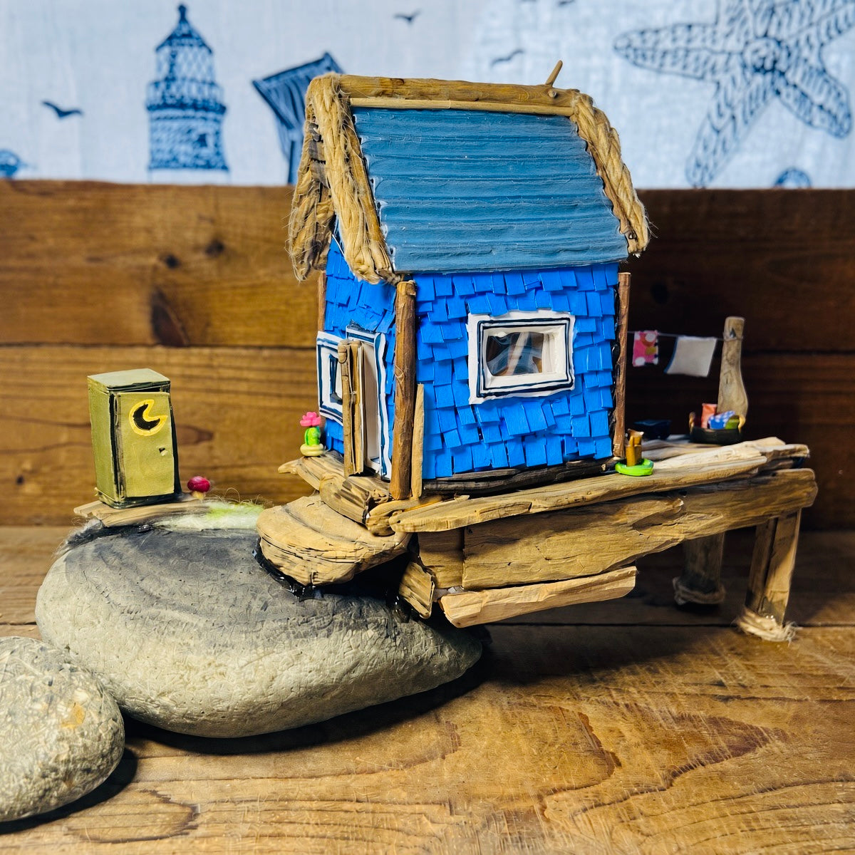 Miniature seaside fishing shack with bright blue shingles, a silver teal roof and driftwood and rope trim, set on a rustic wooden wharf beside grey rocks and a tiny yellow outhouse, with laundry hanging alongside.